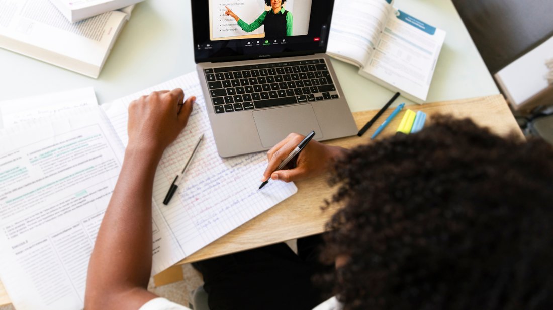 A student on an online math class, writing in a notebook while following along on a laptop